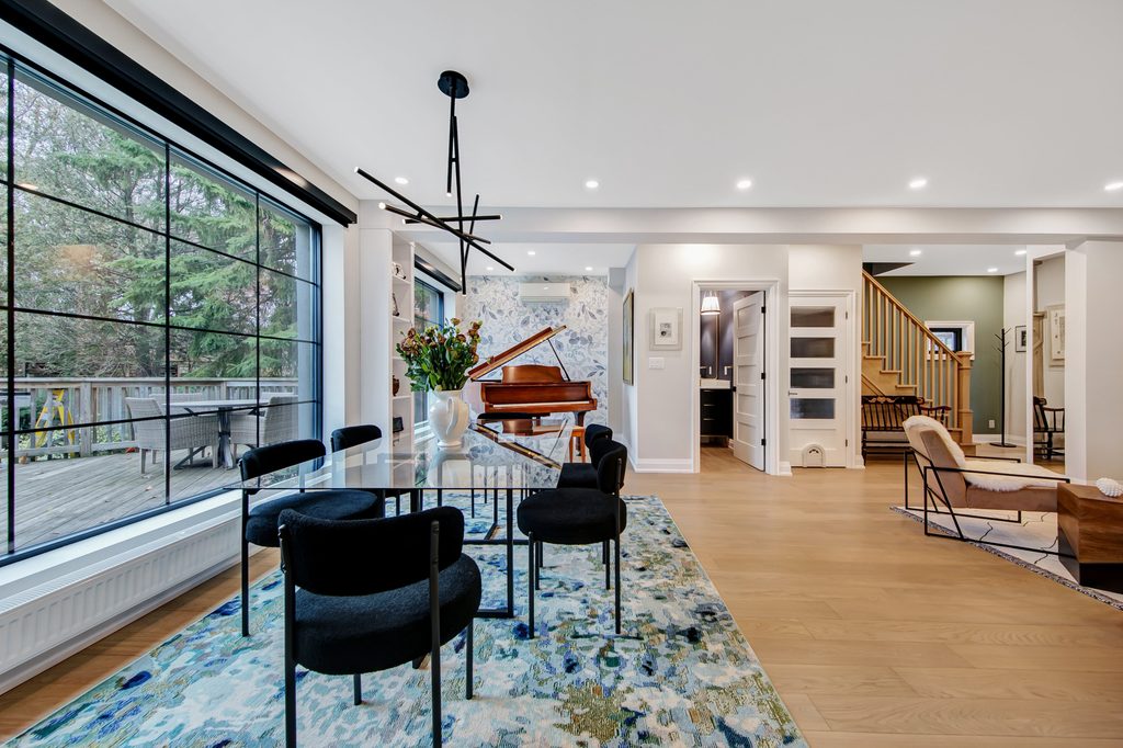 Modern open-plan dining and living area with large window, black chairs, marble table, blue patterned rug, grand piano, wooden staircase, light wood floors, and contemporary lighting.