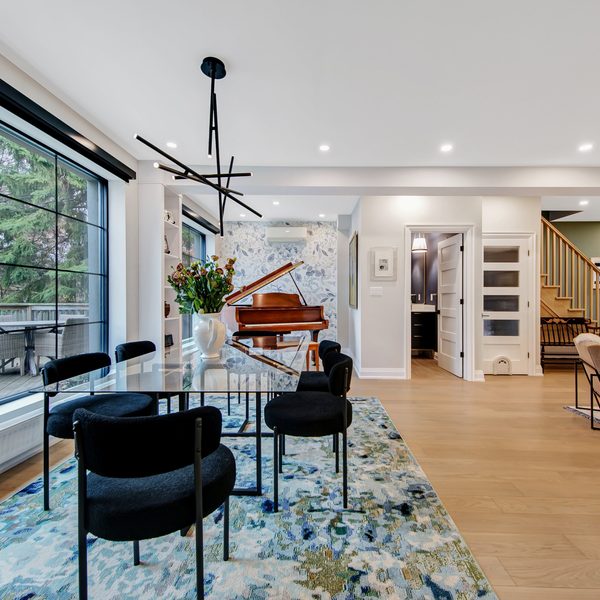 Modern open-plan dining and living area with large window, black chairs, marble table, blue patterned rug, grand piano, wooden staircase, light wood floors, and contemporary lighting.