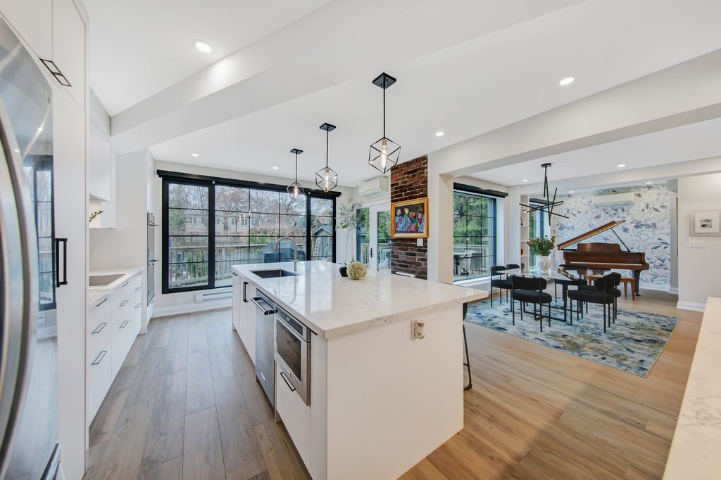 Modern open-concept kitchen with white cabinets, marble island, pendant lights, and wood floors, adjacent to a dining area with a grand piano and large windows overlooking trees.
