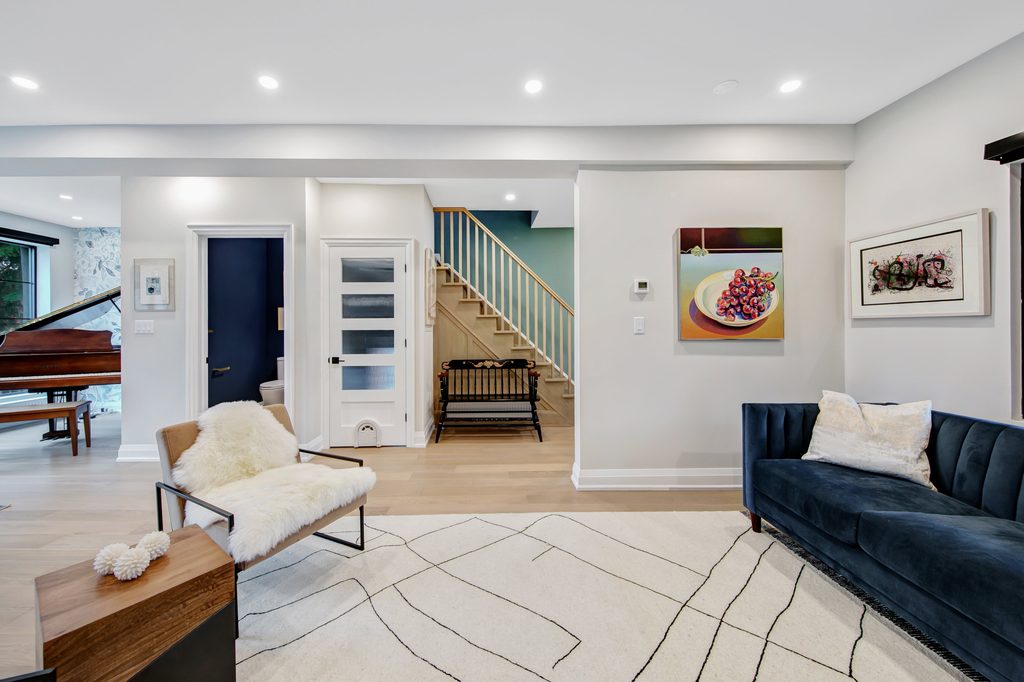 Modern living room with white walls, abstract rug, blue velvet sofa, white fur chair, wall art, and wood staircase. A grand piano is visible in the background, and recessed lighting brightens the space.