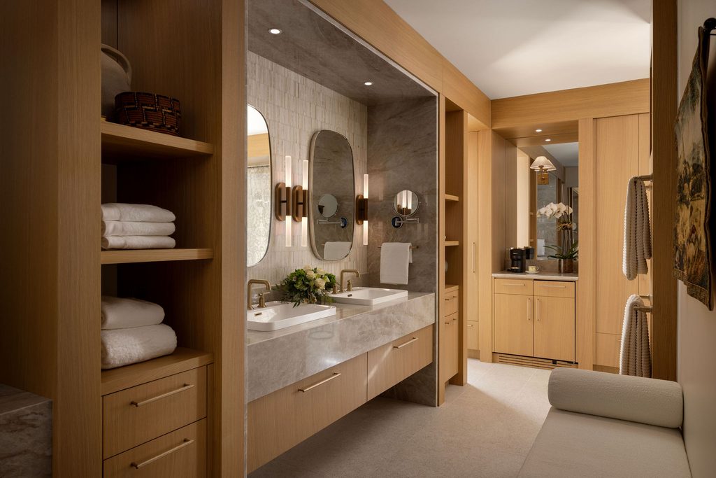 Modern bathroom with double sinks, large mirrors, wooden cabinets, shelves with towels, wall sconces, plants, and a small seating area. A coffee machine is visible on a counter in the background.
