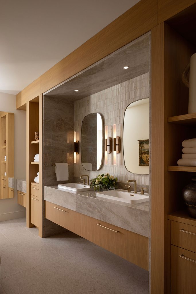 Modern bathroom with a double sink vanity, wood cabinetry, two oval mirrors, wall lights, stacked towels, and decorative plants, creating a warm and elegant atmosphere.