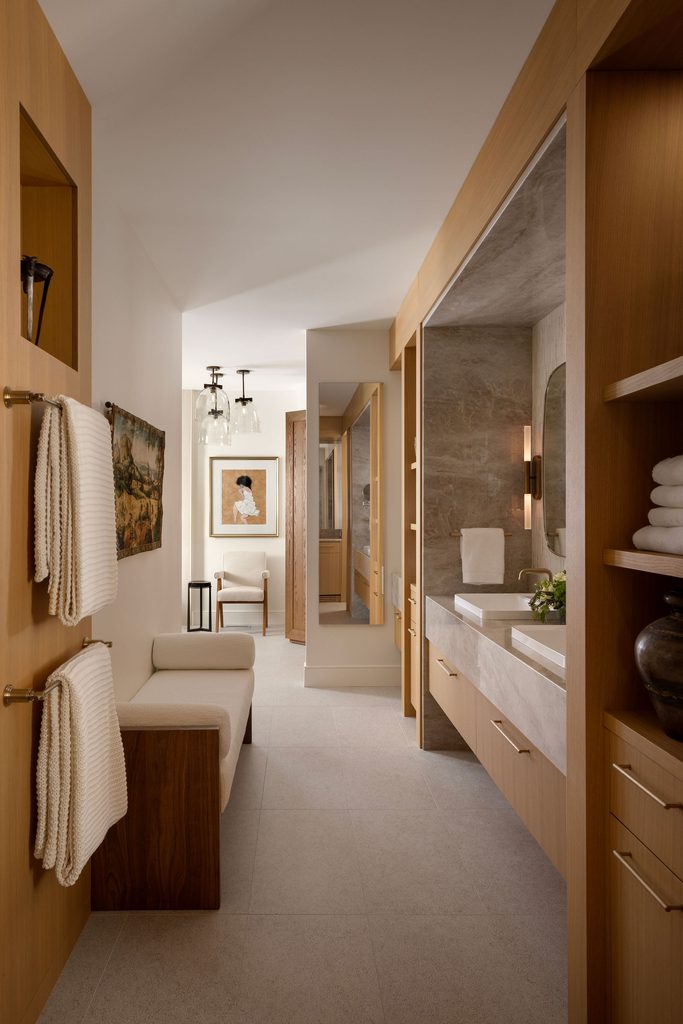 Modern bathroom with wooden accents, white towels on racks, a built-in bench, double sinks with a large mirror, and wall art. The space is well-lit and neatly organized, with light-colored floors and walls.
