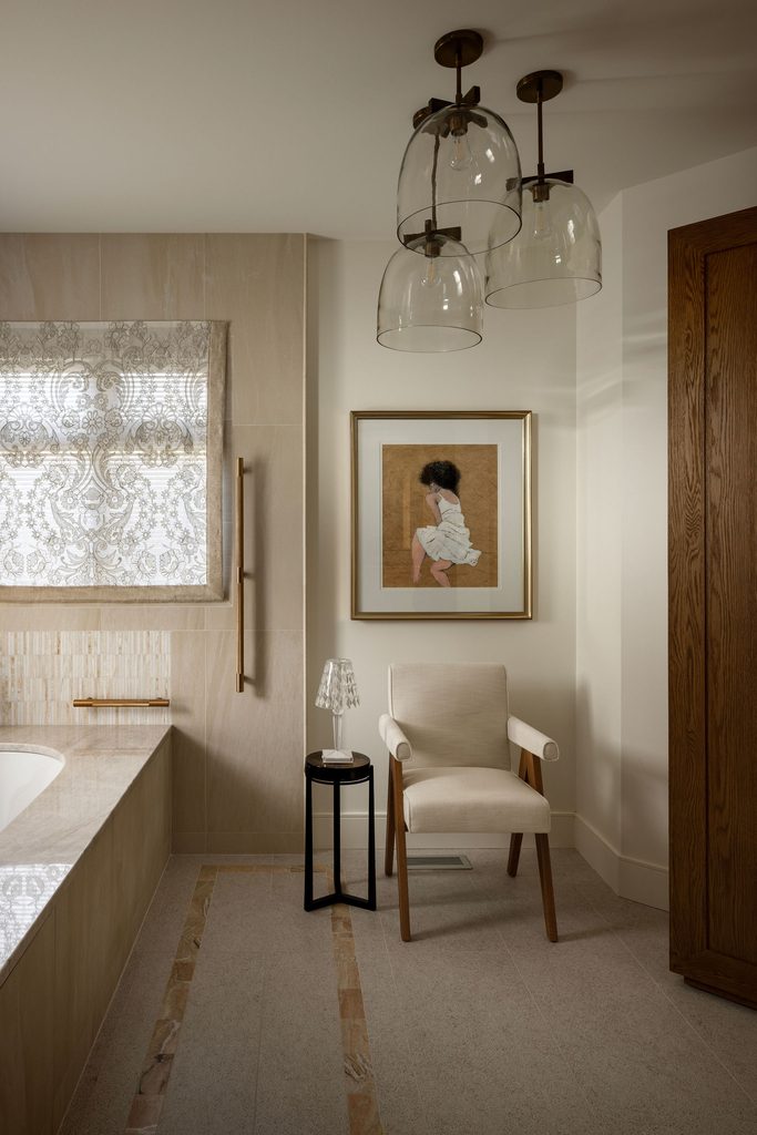 Modern bathroom with neutral tones, a white armchair, small black side table with a glass lamp, framed artwork on the wall, wood accents, and three glass pendant lights hanging from the ceiling.