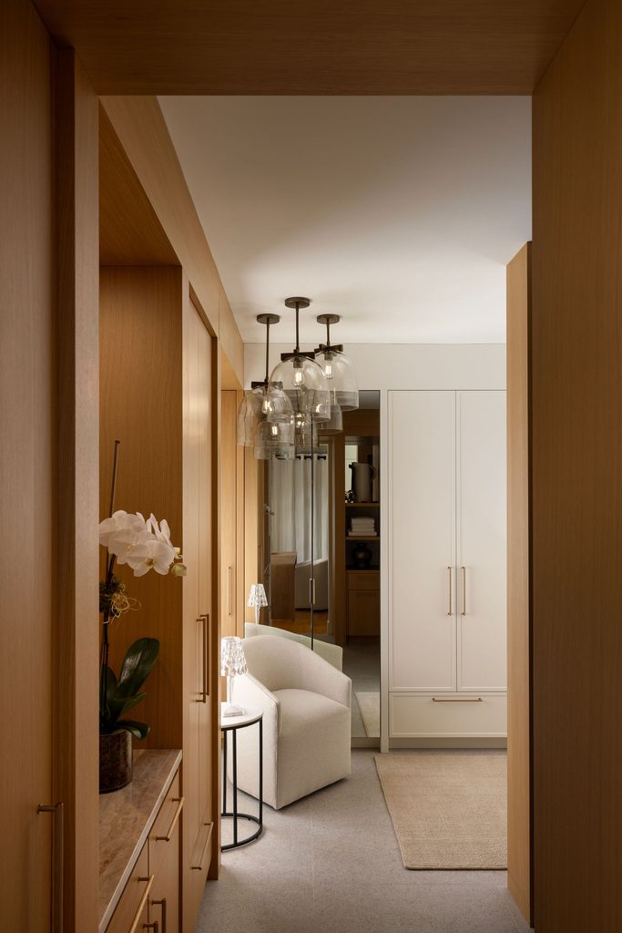 A modern hallway with wood paneling, a white cushioned chair, small side table, hanging glass light fixtures, and a white wardrobe. An orchid sits on a console to the left. A beige rug covers part of the floor.