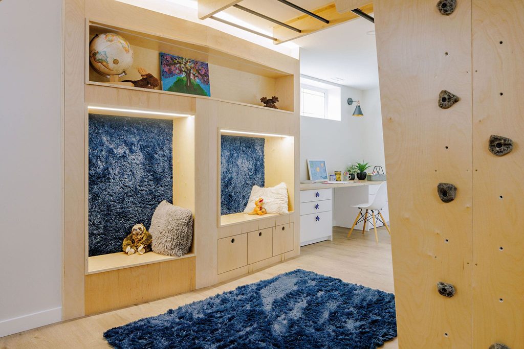 A cozy playroom with built-in wooden seating, blue textured cushions, plush toys, a climbing wall, a blue rug, shelves with books and decor, and a desk with a chair and plant in the background.