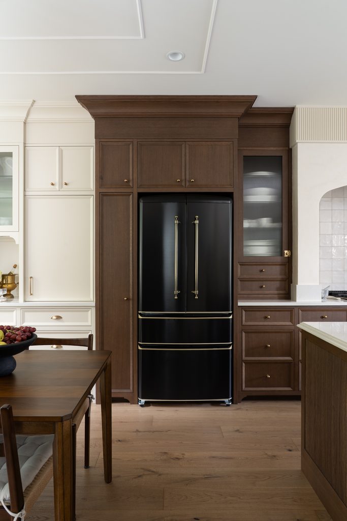 A modern kitchen with wood cabinetry, a black French door refrigerator, white countertops, and a wooden dining table with a bowl of fruit. Light wood flooring and built-in shelves are also visible.