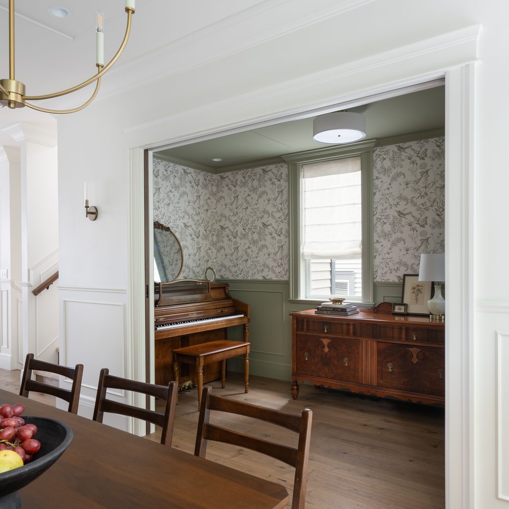 A view into a cozy room with a piano, mirror, and wooden sideboard; floral wallpaper and green wainscoting complement the elegant, softly lit space. Foreground shows part of a dining table and chairs.