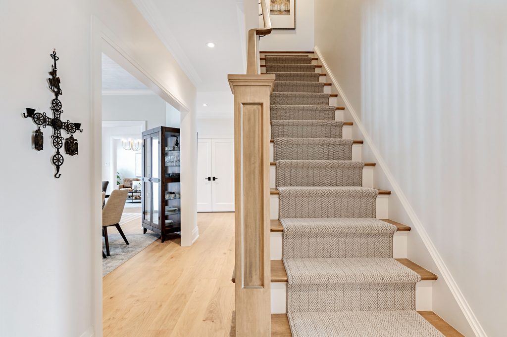 A carpeted wooden staircase with a light wood banister in a bright hallway. To the left is a dining area with a table and chairs. A decorative wall cross hangs beside the staircase.