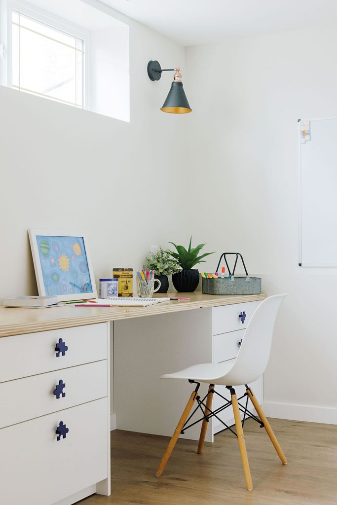 Bright home office with a white desk, modern white chair, plants, art supplies, framed artwork, and a whiteboard on the wall. Light flows in from a small window above.