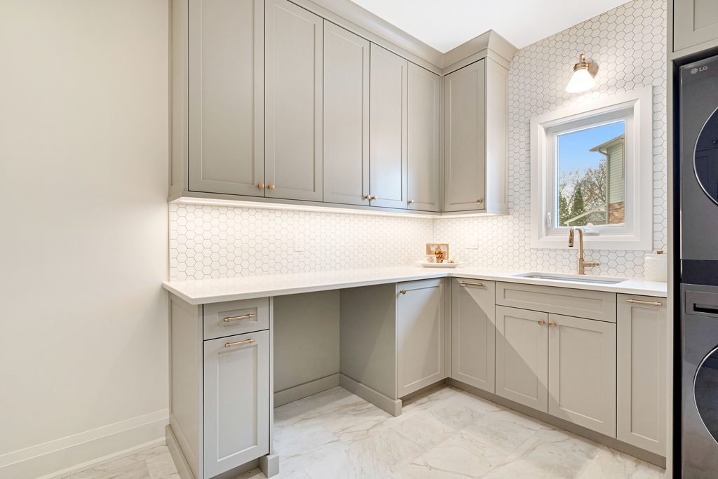 Modern laundry room with gray cabinets, under-cabinet lighting, white hexagon tile backsplash, a small window, stacked washer and dryer, and white countertops. The space is clean and bright.