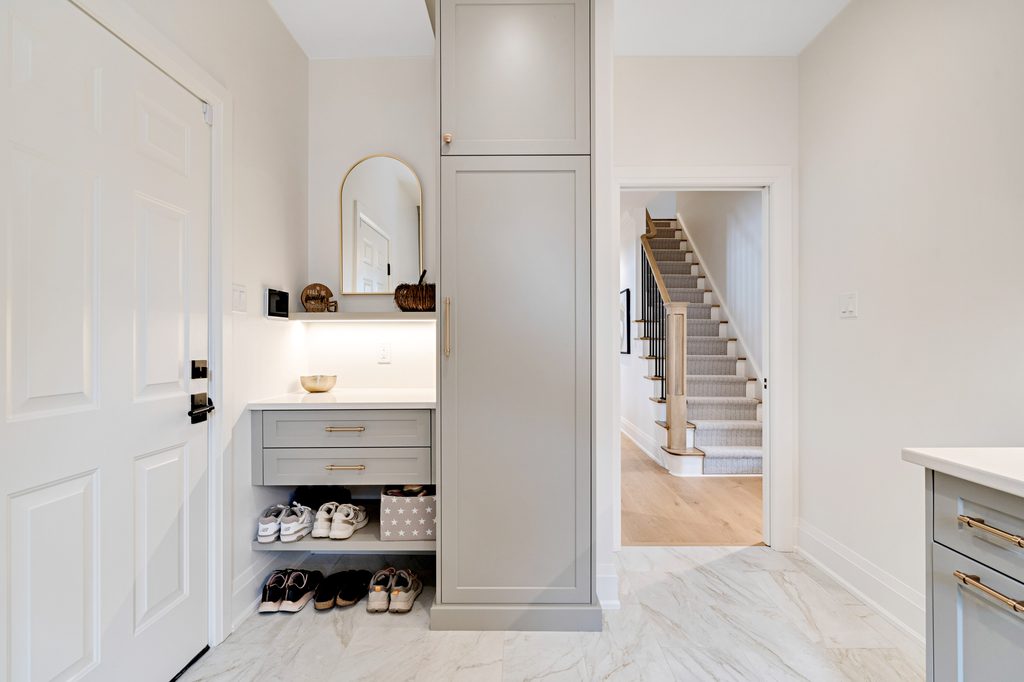 A modern entryway with marble floors, built-in cabinets, a mirror, shelves holding shoes, a white door on the left, and a carpeted staircase leading upward in the background.
