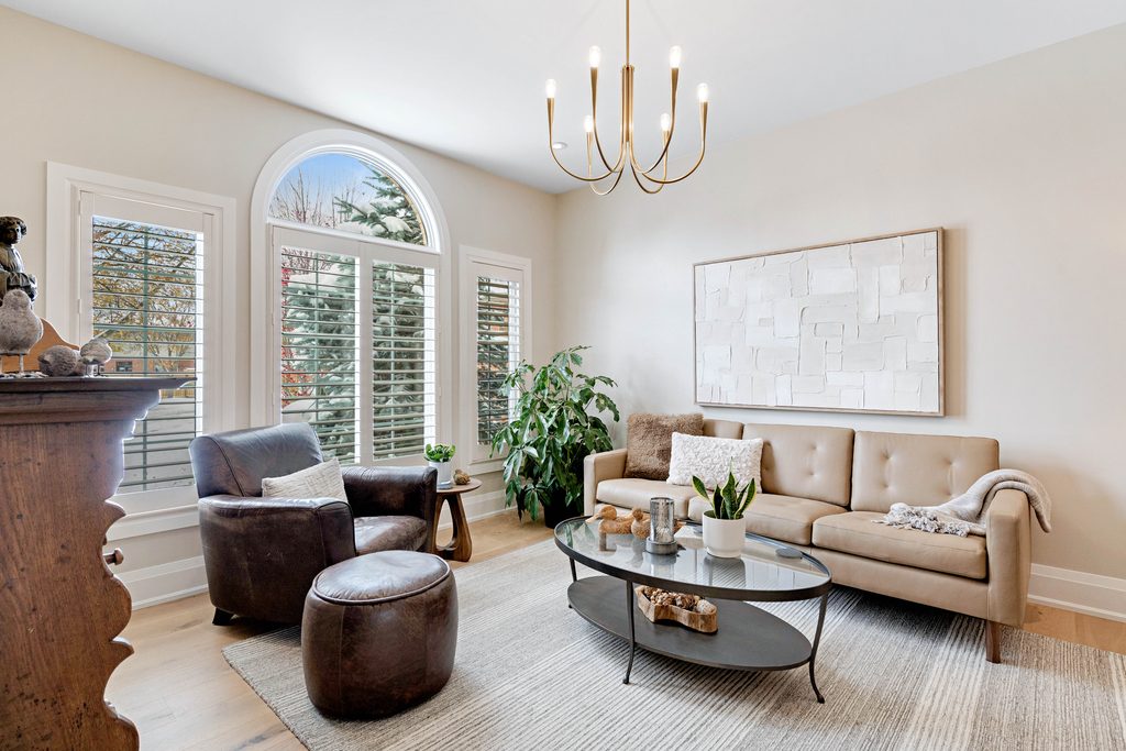 Bright living room with a large arched window, beige sofa, dark leather chair, round coffee table, potted plants, textured wall art, and a modern chandelier. Neutral tones and natural light create a cozy atmosphere.