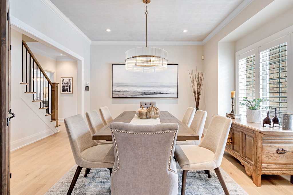 Modern dining room with a long wooden table, eight beige chairs, a large pendant light, wall art, and natural light from windows with shutters. Neutral tones and minimal decor create a bright, elegant space.