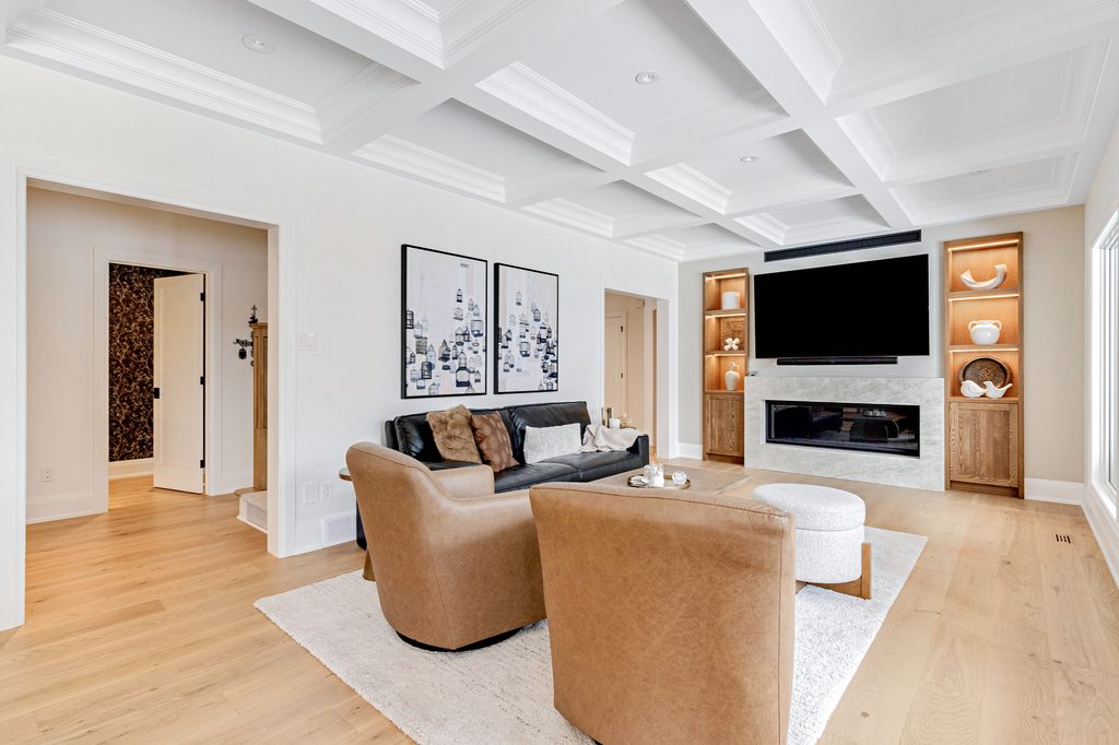 Modern living room with a coffered ceiling, black sofa, two brown armchairs, a round ottoman, built-in shelves, a large TV above a fireplace, and light wood floors. Art and decor pieces are displayed on the walls and shelves.