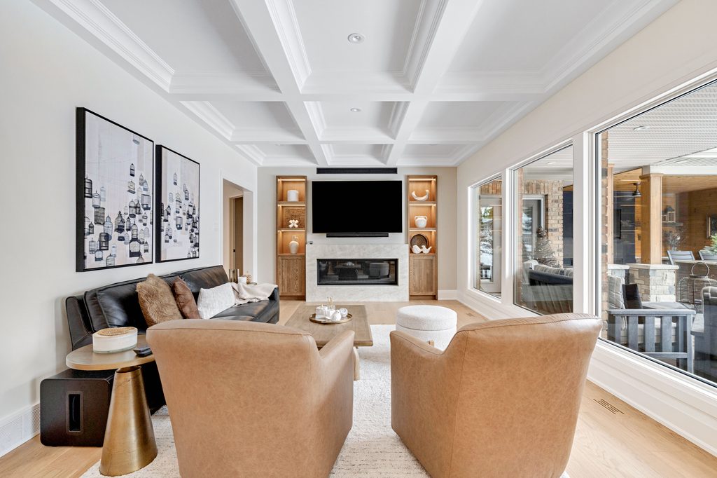 Modern living room with two tan chairs, a black sofa, wall art, a fireplace, a mounted TV, built-in shelves, light wood floors, and large windows showing an outdoor seating area.