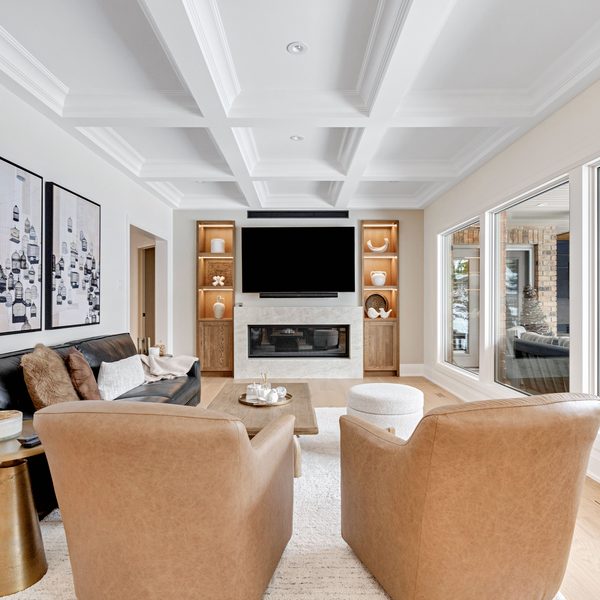 Modern living room with two tan chairs, a black sofa, wall art, a fireplace, a mounted TV, built-in shelves, light wood floors, and large windows showing an outdoor seating area.