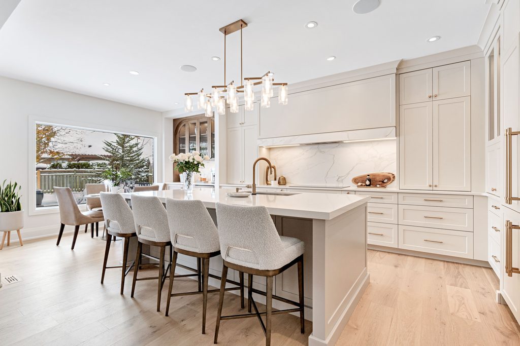 Modern kitchen with a large island, four upholstered chairs, marble countertops, gold fixtures, white cabinets, wood floors, a chandelier, and a window overlooking a yard with trees.