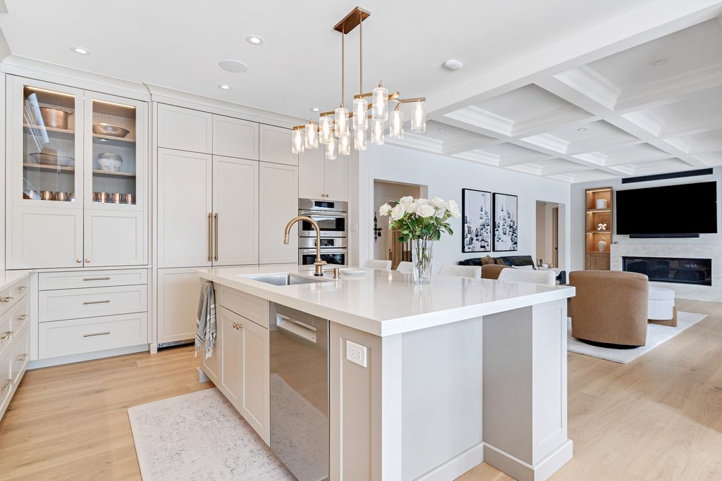 Modern kitchen with white cabinets, a large island with sink, hanging chandelier, and open layout to a bright living room with sofas, art on the wall, and a large TV above a fireplace.