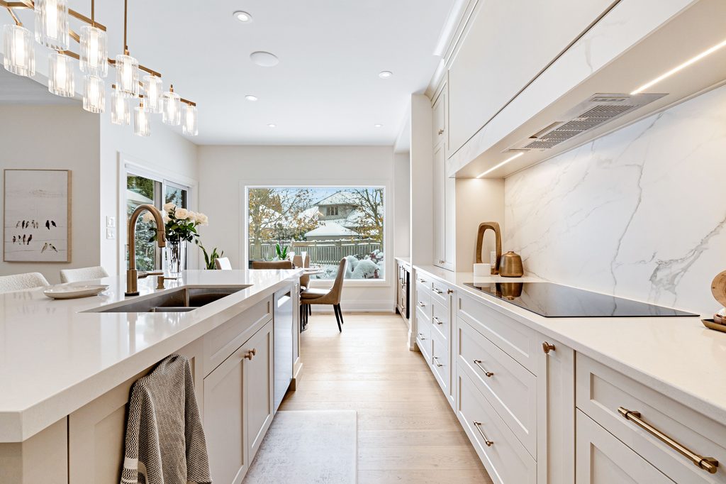 Bright, modern kitchen with white cabinets, marble backsplash, pendant lights, an island with a sink, and large window overlooking a backyard. Dining area visible in the background.
