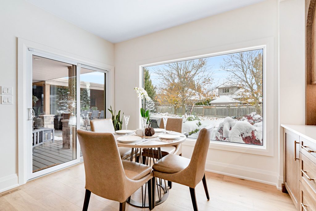 A modern dining area with four beige chairs around a round table, large window showing a snowy backyard, and sliding glass doors leading to a covered patio.