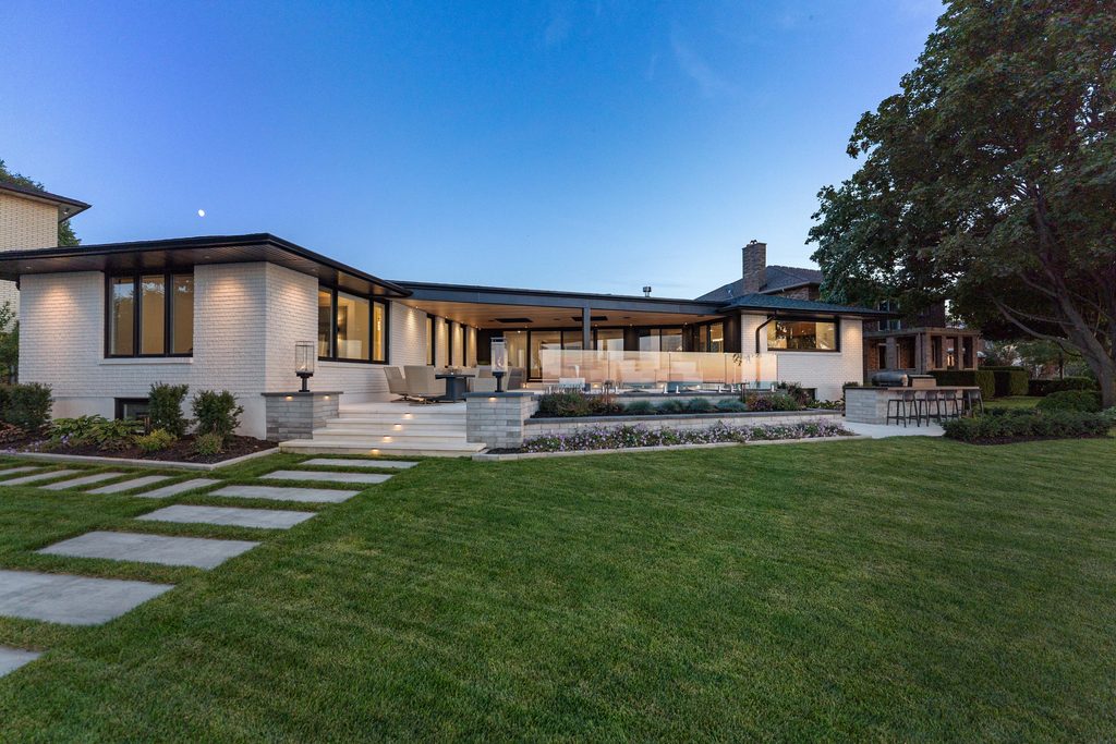 Modern single-story house with large windows, patio seating, and a swimming pool in the backyard, surrounded by green grass, trees, and stone walkway at dusk.