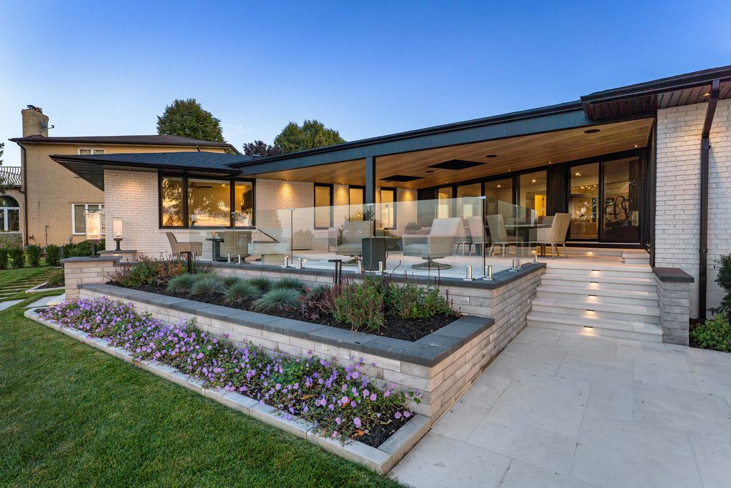 Modern house with large windows, a covered patio with seating, landscaped garden beds, and wide steps leading to a green lawn, shown at dusk under a clear sky.