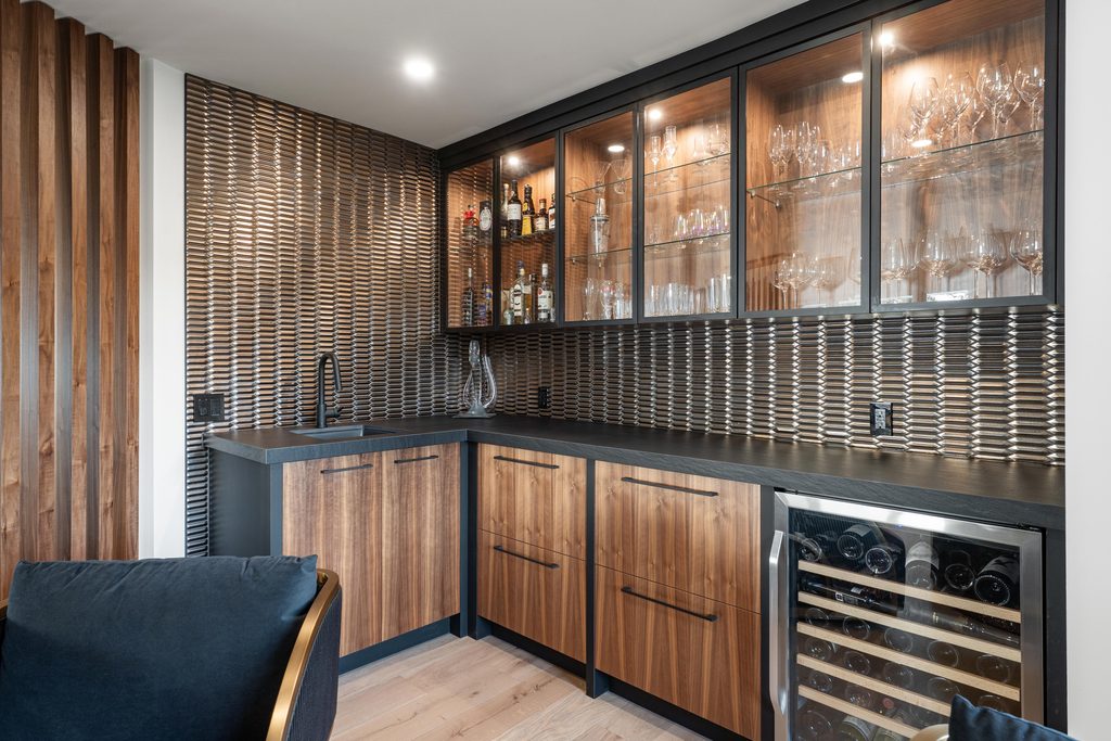 Modern home bar with wood cabinets, black countertops, glass-front upper cabinets displaying glassware, a small sink, wine fridge, and a textured brown wall, with wooden slats on the left and a dark chair partially visible.