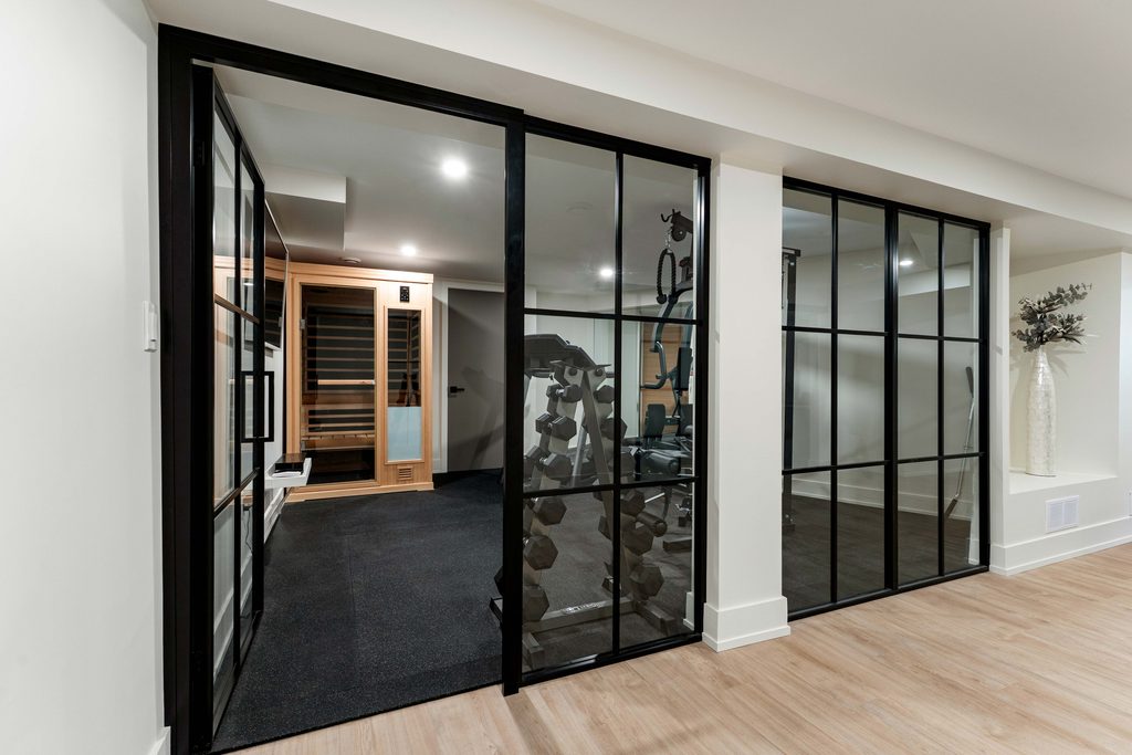 Home gym with glass-paneled walls, a weight rack, exercise equipment, and a wooden sauna; light wood flooring and a decorative vase in the hallway.