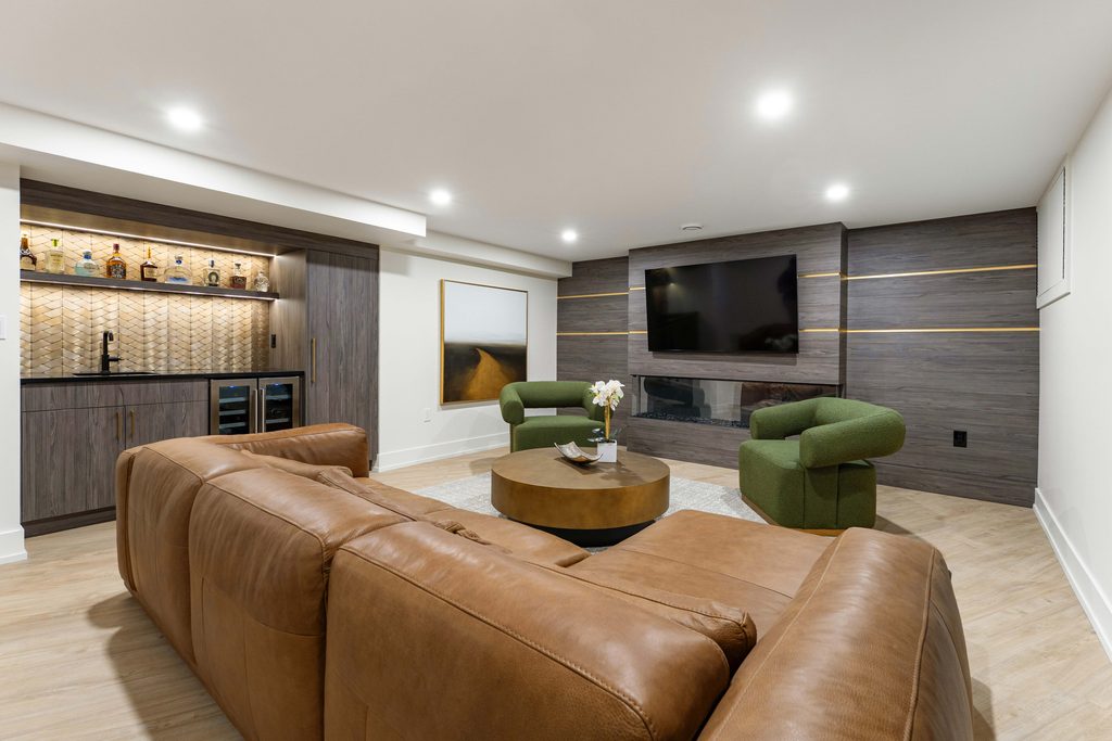 Modern basement living room with a brown leather sofa, two green armchairs, a round coffee table, a wall-mounted TV, a fireplace, and a built-in bar area with shelves, cabinets, and a mini fridge.