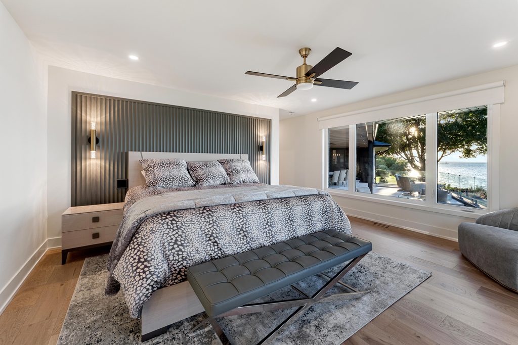 Modern bedroom with a patterned bed, gray bench, ceiling fan, and large window overlooking a patio and water view. Light wood floors and minimal decor create a clean, relaxing atmosphere.