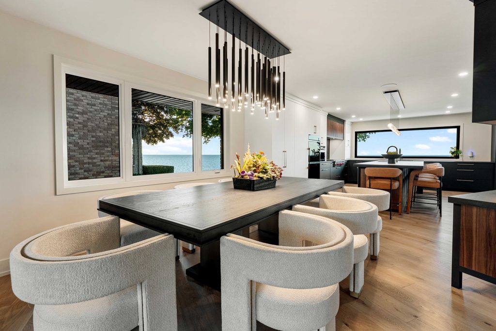 Modern dining room with a black rectangular table, beige cushioned chairs, a flower centerpiece, large windows showing a lake view, and a black linear chandelier above the table. Kitchen with island and barstools in the background.
