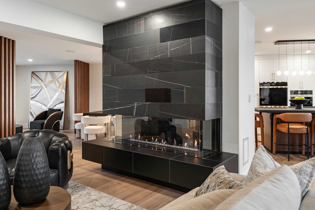 Modern living space featuring a double-sided black tile fireplace, with seating areas on both sides, warm lighting, and an open kitchen visible in the background.