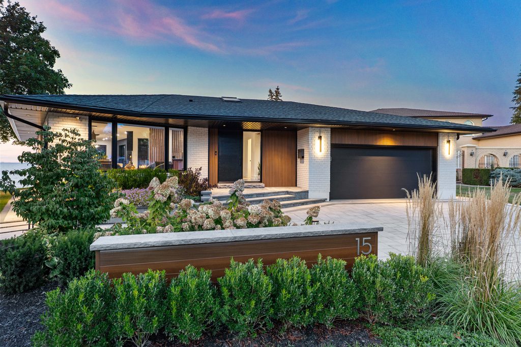 Modern single-story house with white brick, wood accents, large black garage door, landscaped front yard, and a sign showing the number 15, at sunset.