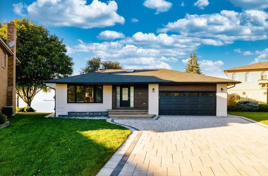 A modern single-story house with a dark roof, light brick exterior, large windows, and a double garage, set against a blue sky with scattered clouds and surrounded by green lawn and trees.