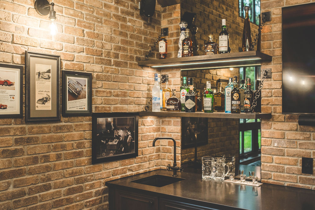 A small home bar with a brick wall, framed photos, shelves of liquor bottles, a black sink, glassware on the counter, and a mirror reflecting the room and window.