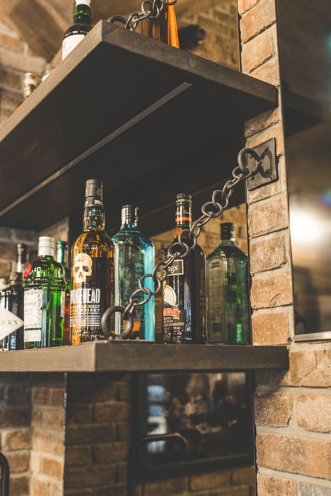 Various liquor bottles are displayed on a wooden shelf with metal chains, attached to a brick wall. The scene is indoors with a rustic, cozy atmosphere.