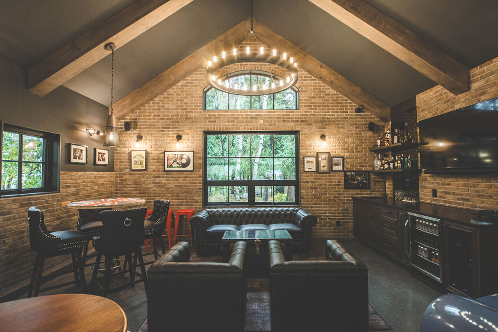 A cozy lounge with leather sofas, brick walls, large windows, a circular chandelier, bar area, and framed photos, blending rustic and modern styles.