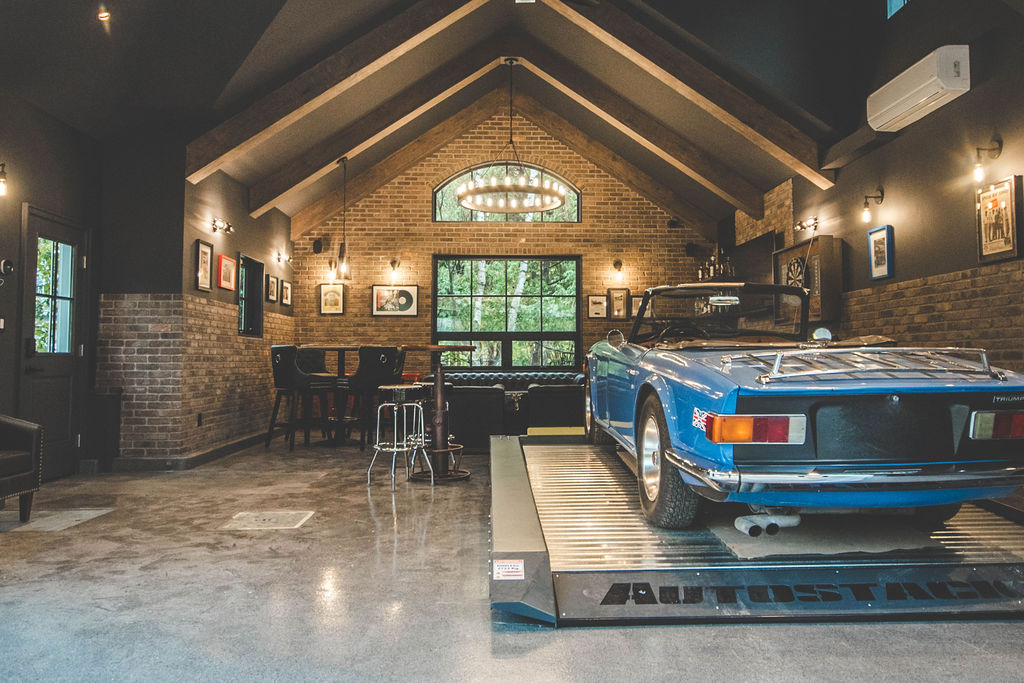 A blue classic convertible car is parked on a lift in a stylish garage with brick walls, art on display, a bar area, and large windows providing natural light.