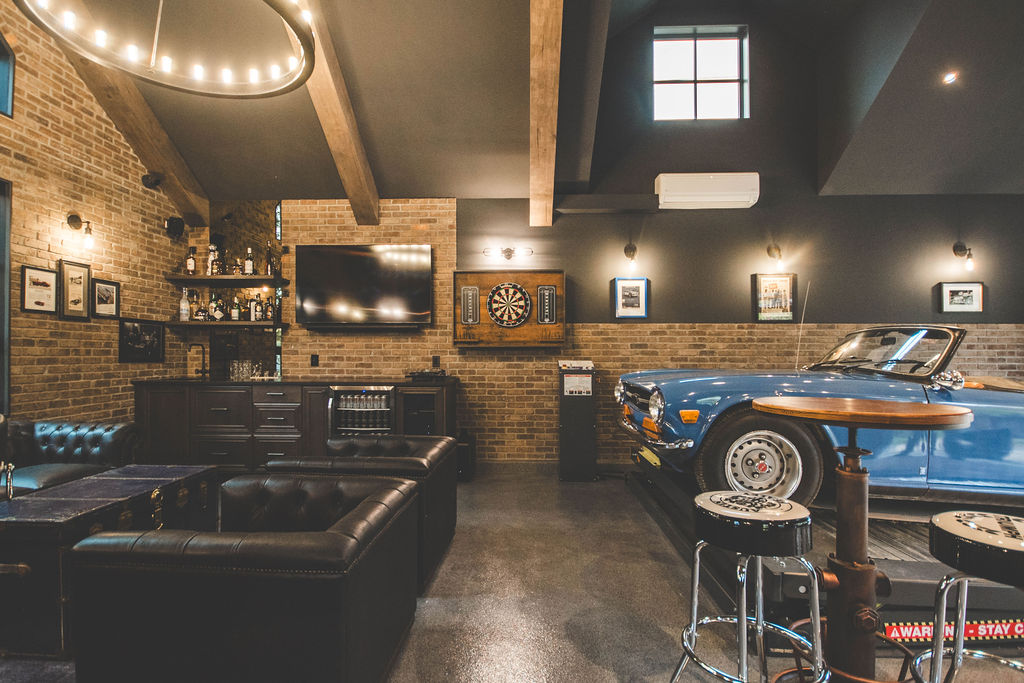 A cozy lounge with leather couches, a TV, a dartboard, exposed brick walls, a vintage bar, and a classic blue convertible parked indoors. Framed photos and drinks decorate the space.