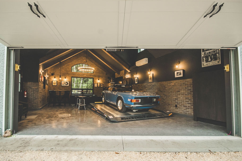 A blue vintage car is parked on a platform inside a stylish garage with brick walls, modern lighting, and classic decor, viewed through an open garage door.