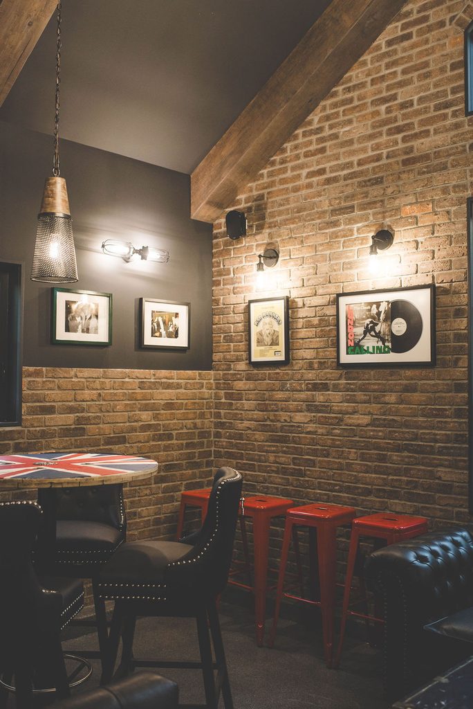 A cozy room with exposed brick walls, black leather chairs, red metal stools, a round table, hanging lights, and framed art and records on the wall.