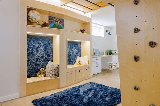 Modern kids playroom with built-in cozy reading nooks, plush blue rug, pillows, stuffed animals, wall shelves, and a climbing wall. A small desk with a chair and plants is in the background.
