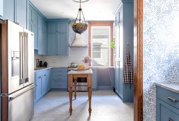 A bright kitchen with blue cabinets, a center island, stainless steel fridge, hanging basket with fruit, and a large window letting in natural light. A checked apron hangs by the window.
