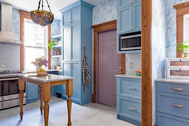 A kitchen with blue cabinets, wood trim, a purple door, a stained glass pendant light, and a small wooden island. Appliances and flowers sit on counters, with a checkered apron hanging by the door.