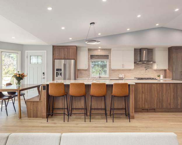 Modern kitchen with wood accents, white cabinets, stainless steel appliances, four brown barstools at an island, a dining table with flowers, and large windows letting in natural light.