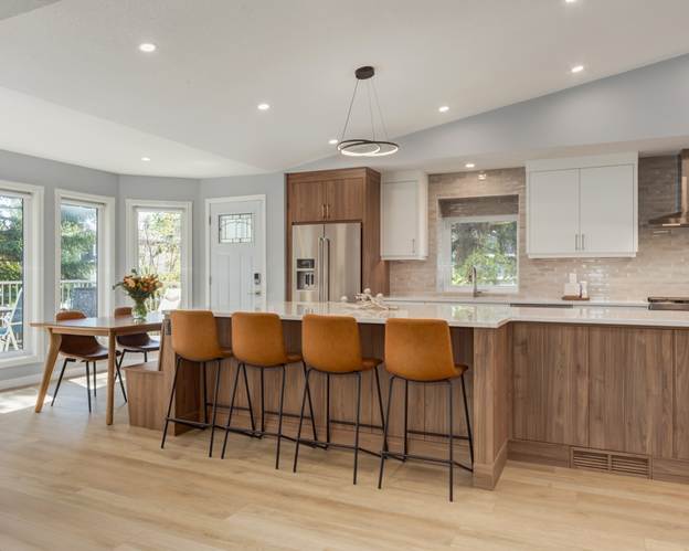 Modern kitchen with wood floors, large island with four brown barstools, stainless steel appliances, white and wood cabinets, pendant lighting, and dining table with flowers by bright windows.