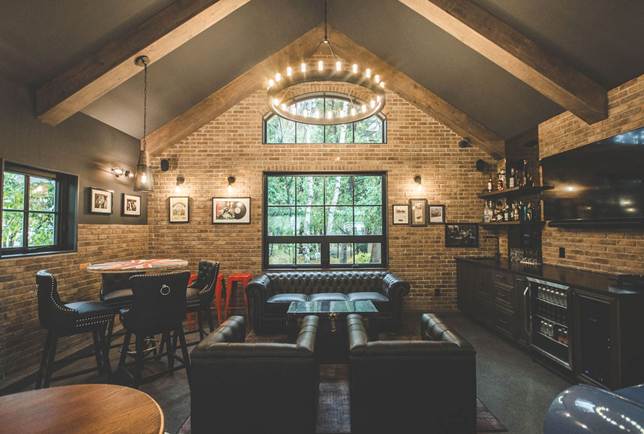 Cozy lounge with brick walls, large windows, leather chairs, a round chandelier, bar area, and framed art. Exposed beams and rustic decor create a warm, inviting atmosphere.