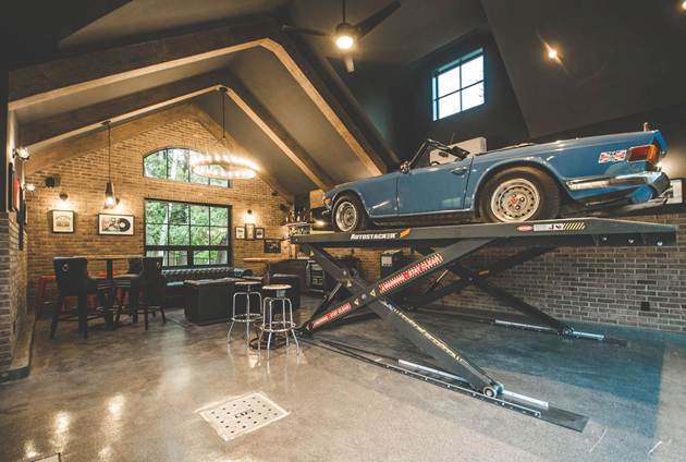 A vintage blue convertible car is elevated on a hydraulic lift in a stylish garage with brick walls, bar seating, framed photos, and large windows letting in natural light.