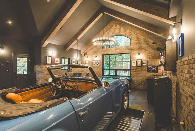 A vintage blue convertible is parked inside a stylish garage with exposed brick walls, large windows, wooden beams, and framed pictures on the walls. Warm lighting creates a cozy, upscale atmosphere.